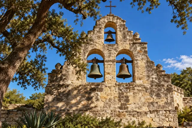 A stone mission bell wall stands beneath oak branches against a blue sky.